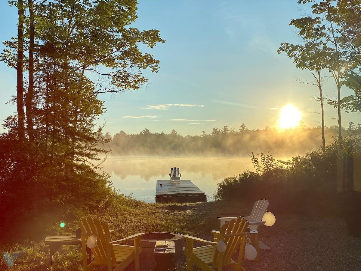 A tranquil lakeside view showcases a private dock surrounded by mist, with the sun rising over the horizon. Two Adirondack chairs sit in the foreground, arranged around a fire pit, creating an inviting space for relaxation and contemplation.