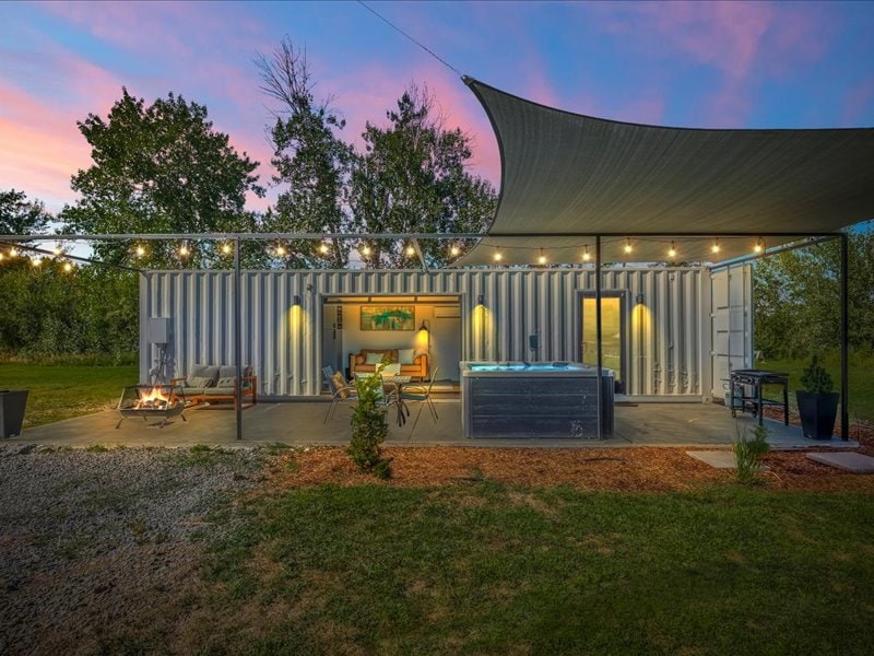 An outdoor view of the container home reveals a cozy seating area under string lights, with a hot tub situated nearby. A shaded patio extends to the open landscape, surrounded by greenery. The twilight sky adds warmth to the inviting atmosphere, creating a peaceful retreat.
