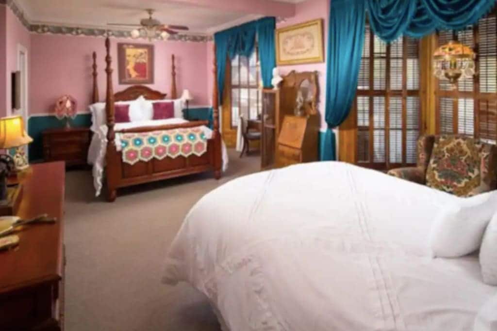 The spacious room features a queen bed and a double bed, each adorned with distinct bedspreads. A seating area is established with an upholstered chair, complementing the elegant decor characterized by pink walls and blue drapery, alongside wooden furnishings and privacy-enhancing window shutters.