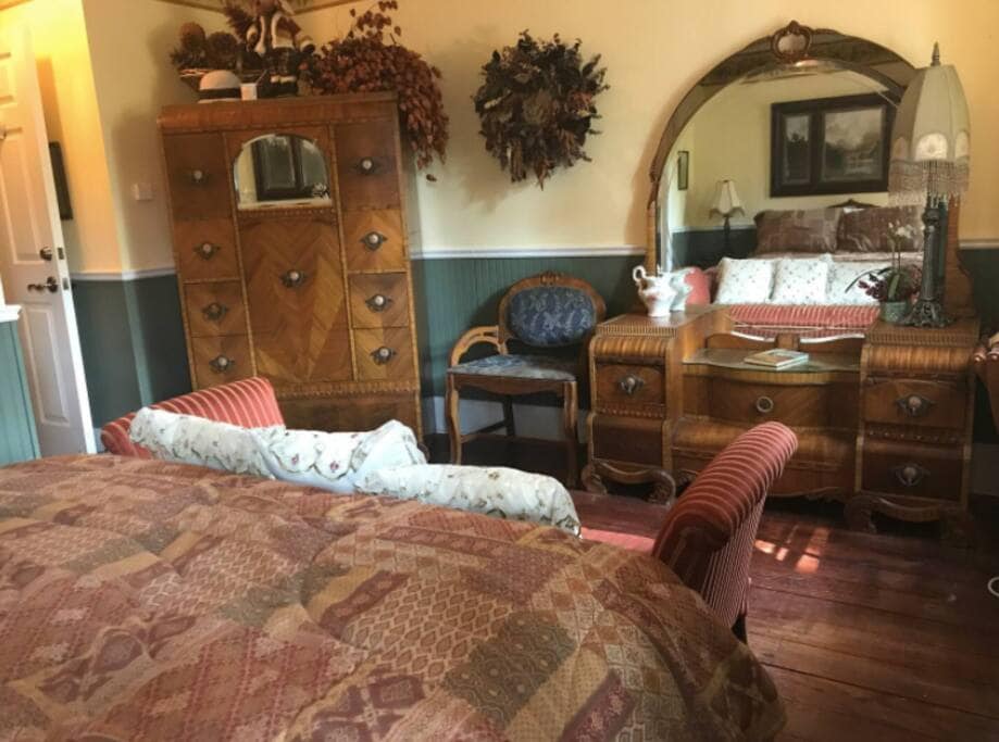 A cozy sleeping area is highlighted by a patterned bedspread covering a spacious bed. Antique furniture, including a dresser with a mirror and a decorative armchair, complements the room. Warm colors and wooden floors add to the room's inviting look.