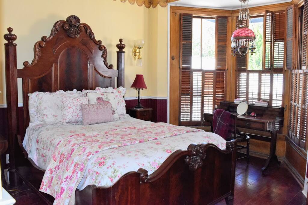 The room features a queen bed with a floral patterned quilt and decorative pillows. Natural light filters through wooden shutters and a bay window. A vintage writing desk is set near the window, complemented by a lamp and rich wood flooring.