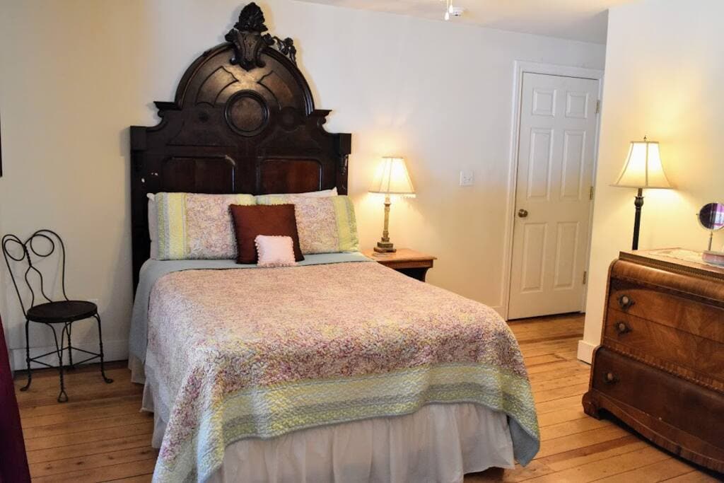 A cozy sleeping area is presented with a full size bed adorned in a patterned bedspread. Two lamps flank the bed, providing soft illumination. A wooden dresser and a small chair are visible, complemented by warm wood flooring and a door leading to a shared bathroom.