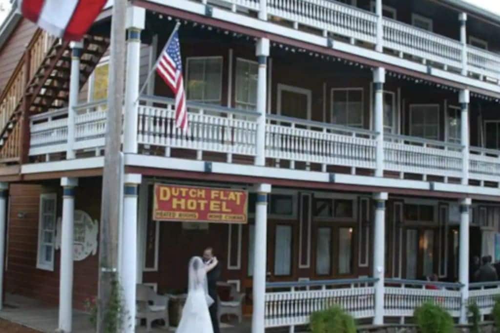 The Historic Dutch Flat Hotel features a charming facade with multiple balconies and white railings. An American flag hangs prominently, adding to the building's historic character. The entrance is framed by a welcoming porch area with seating, surrounded by well-maintained greenery.