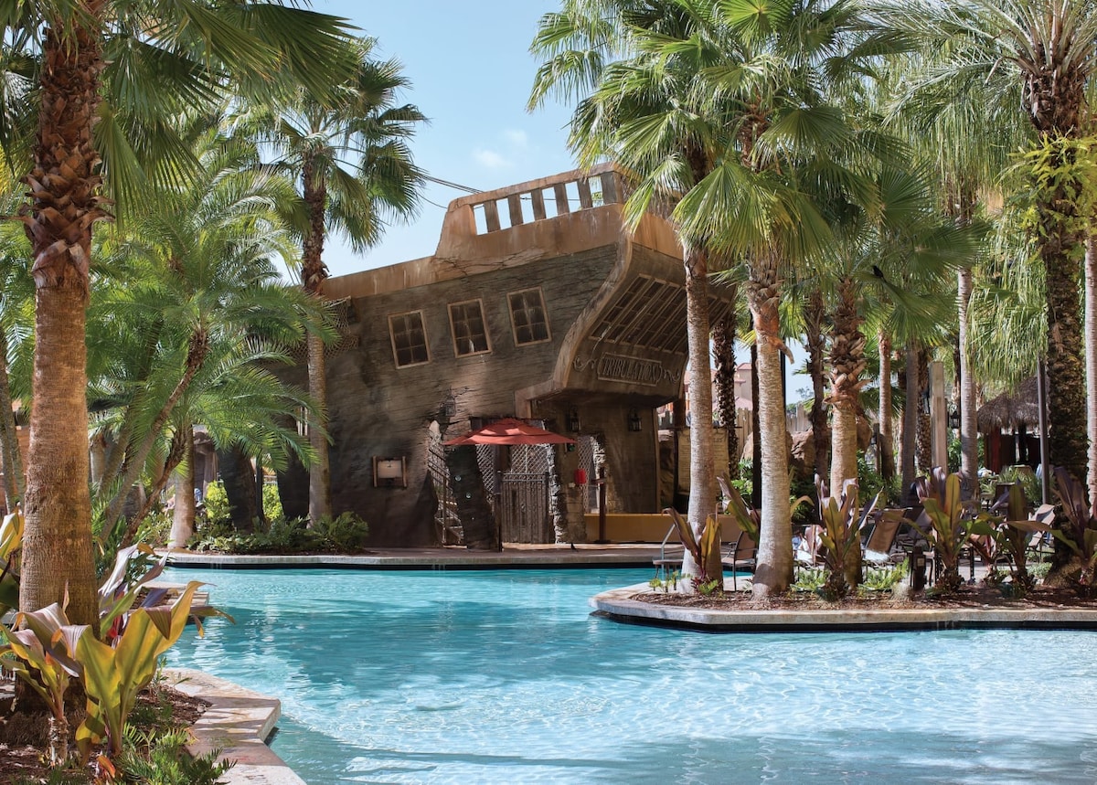 A large, inviting pool is surrounded by lush palm trees and tropical plants, creating a relaxing oasis. A shaded structure with seating is visible in the background, offering a cool reprieve from the sun. Clear blue water reflects the bright sky.