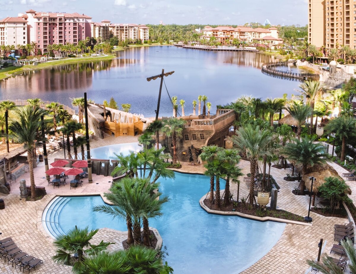 A lagoon-style pool is surrounded by palm trees, with a pirate ship play area in view. Red patio umbrellas and lounge chairs are situated around the pool, while a serene lake reflects the surrounding buildings and lush greenery.