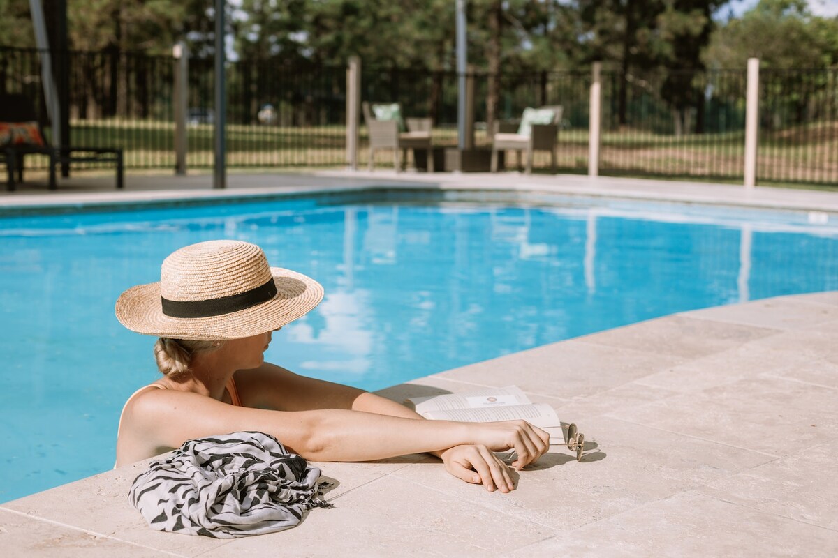 A pool area features a clear blue water and a surrounding patio. A person is relaxing by the edge, wearing a straw hat and holding a book. Deck chairs and a grassy area are visible in the background, enhancing the tranquil setting.