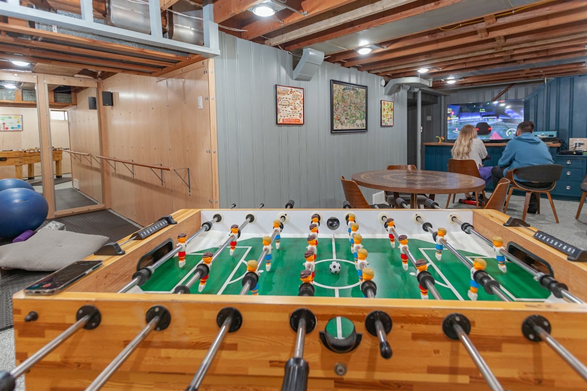 A vintage foosball table occupies the foreground, featuring colorful player figures on a green field. In the background, a cozy seating area with a television is visible, where two people sit together. The space includes gym equipment and cabinets against the walls, enhancing a communal atmosphere.