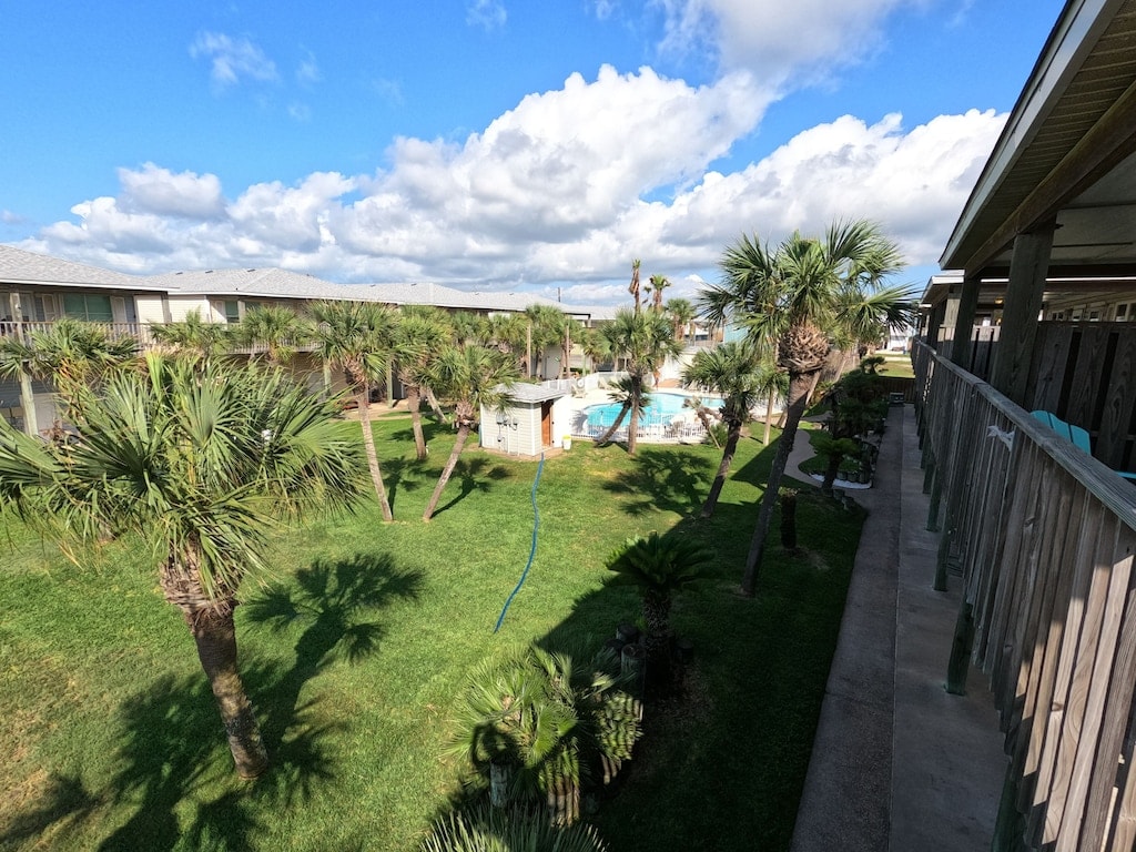 A landscaped outdoor area is visible, featuring lush green grass and palm trees. The scene includes a pool surrounded by a spacious deck and a building with multiple entrances. The sky is partly cloudy, providing a bright atmosphere across the landscape.