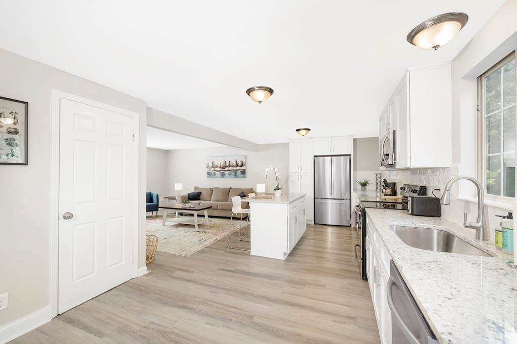 An open kitchen showcases granite countertops and stainless steel appliances, including a refrigerator and oven. Ample cabinetry provides storage space. The adjacent living area is visible, featuring comfortable seating and a decorative rug, creating a seamless flow between cooking and gathering spaces.