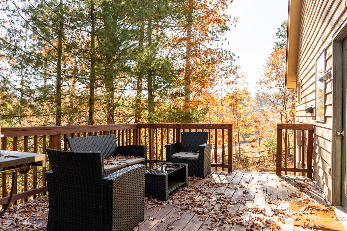 A spacious outdoor deck features a comfortable seating arrangement with dark wicker furniture and a coffee table. The surrounding trees showcase autumn foliage, enhancing the serene atmosphere. A grill is visible in the corner, making it ideal for outdoor cooking.