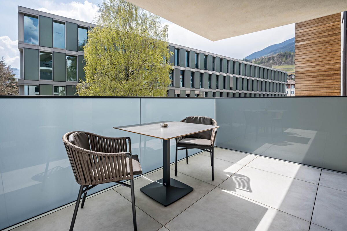A spacious private balcony features a rectangular table with a centerpiece, surrounded by two comfortable, woven chairs. Large glass panels provide a clear view of the outdoors, where greenery and modern architecture can be seen in the backdrop.
