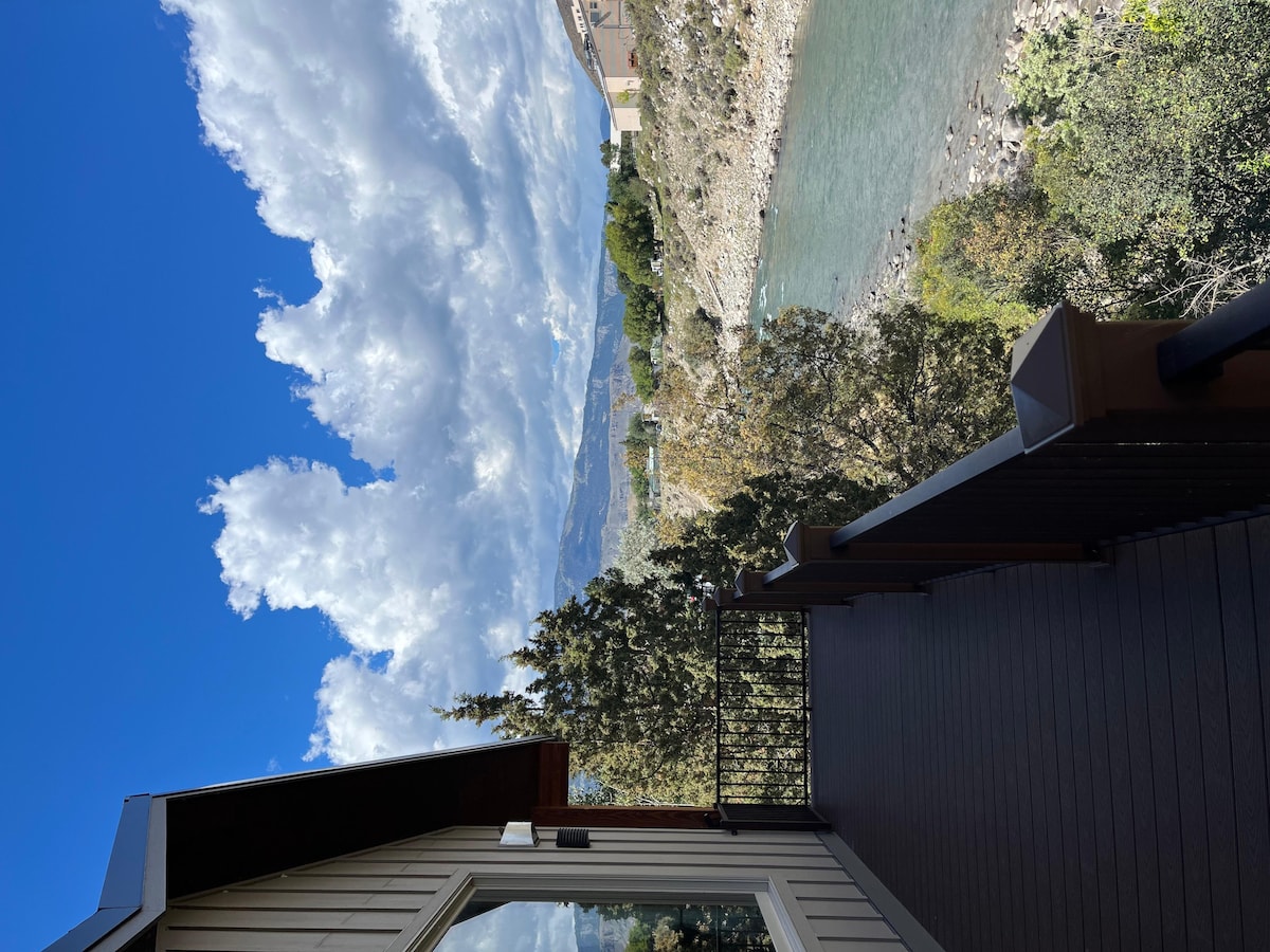 A private deck provides a view of the Yellowstone River, framed by trees and mountains in the background. Bright blue skies and fluffy white clouds enhance the natural scenery, creating a peaceful outdoor space for relaxation and wildlife observation.
