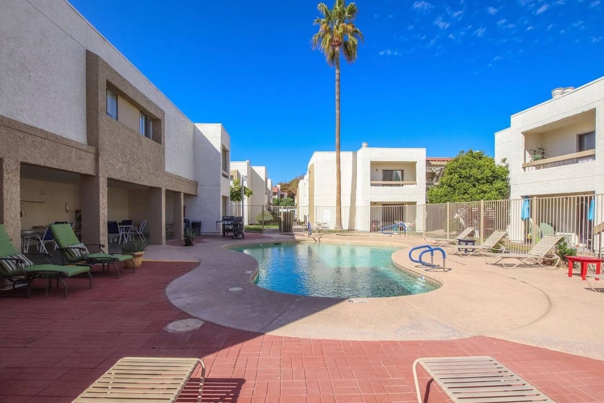 A serene community pool is surrounded by lounge chairs and palm trees, with the sun illuminating the water. The area is framed by well-maintained, white stucco buildings that provide a relaxing backdrop, creating a welcoming space for leisure and enjoyment.
