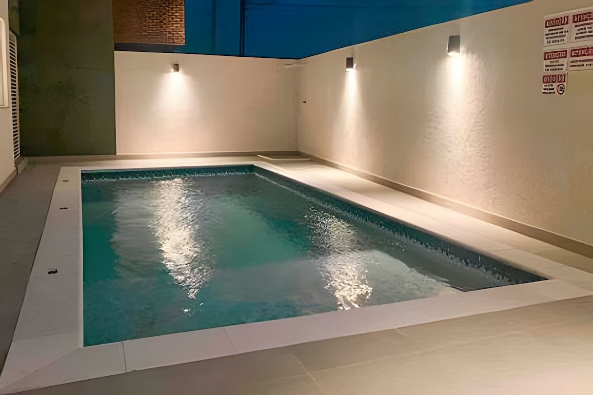 A clean and modern outdoor swimming pool is illuminated by soft wall-mounted lights, reflecting the calm water. The pool area is surrounded by sleek, tiled surfaces, offering a relaxing environment for guests.