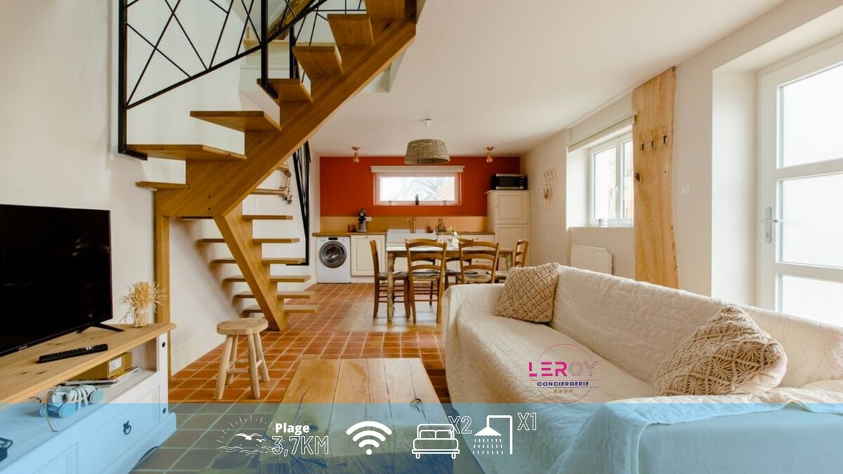 An open living area features a light-colored couch facing a television unit. A staircase leading to the upper floor is highlighted by an intricate railing. A dining table surrounded by chairs is visible in the background, with a washing machine discreetly placed to one side.