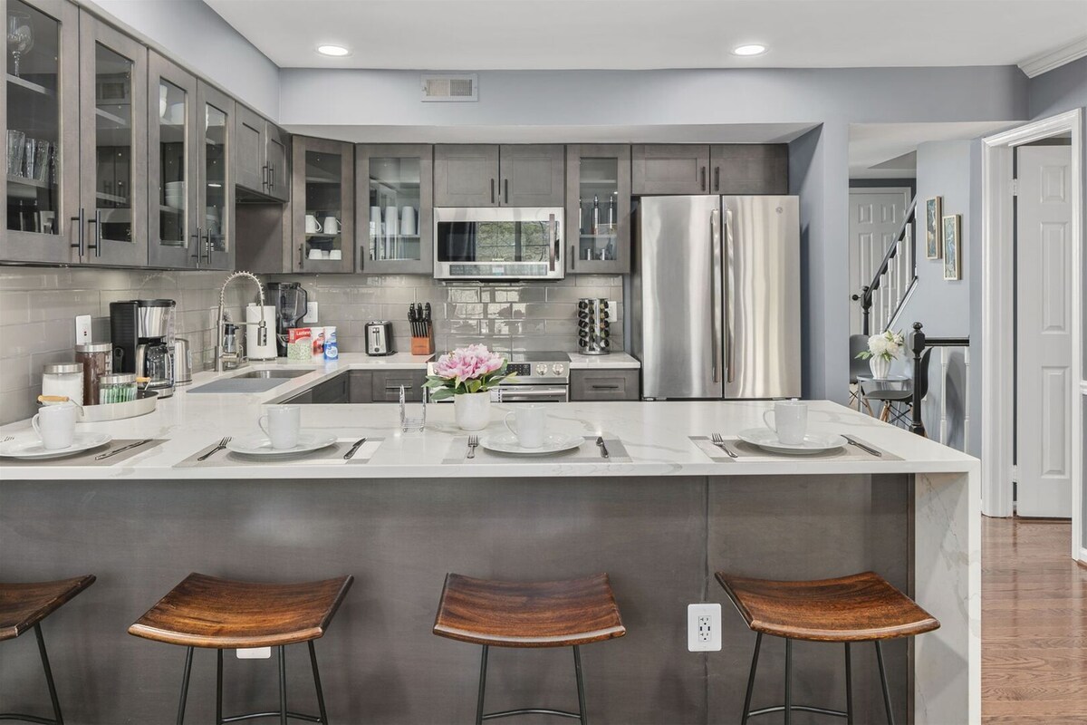 The modern kitchen features matte black cabinets complemented by sleek Carerra marble countertops with a waterfall edge. Stainless steel appliances are visible, along with elegant dishware arranged neatly. The kitchen island includes four wooden stools, creating an inviting space for dining or socializing.