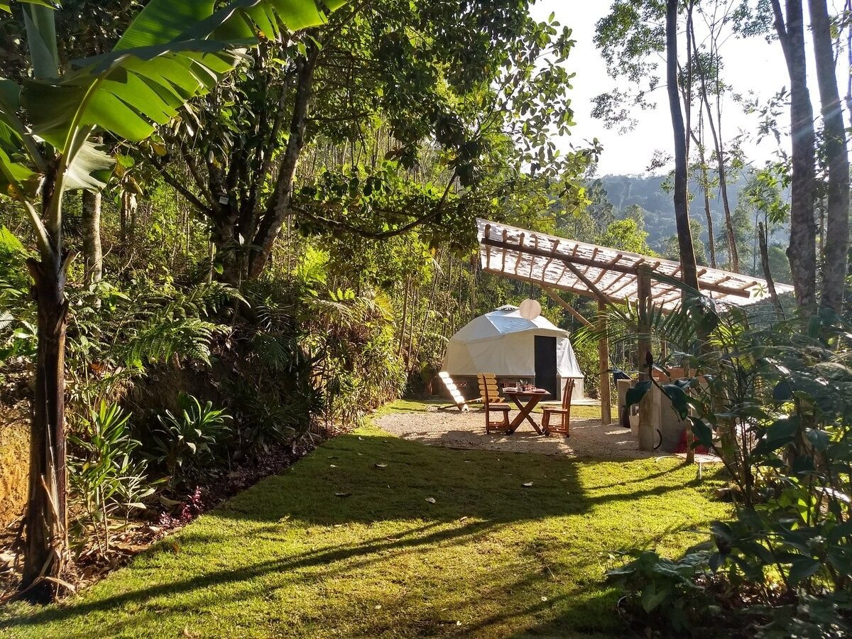 A bright outdoor space is surrounded by lush greenery, with a white geodesic dome positioned on a clear patch of grass. Nearby, a wooden table with benches is set beneath a rustic roof structure, creating a cozy spot for relaxation and connection with nature.