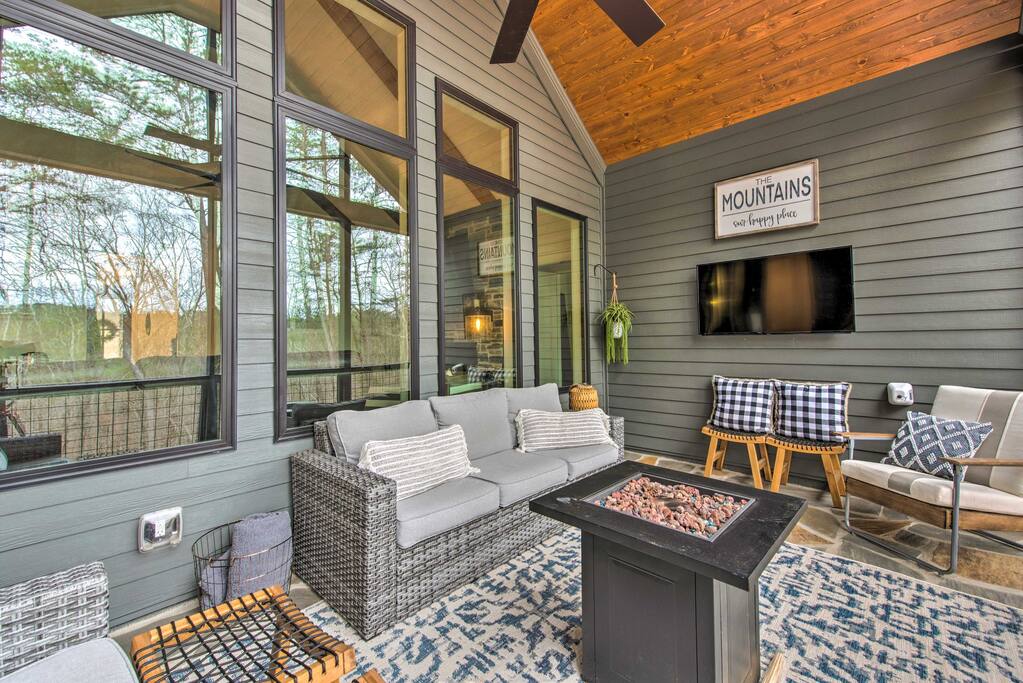 A screened-in patio area is furnished with a comfortable seating arrangement, featuring a cozy gray sofa and an outdoor armchair beside a fire table. Large windows provide views of the surrounding nature, and a mounted TV adds entertainment options. A decorative wall sign reads 'MOUNTAINS.'