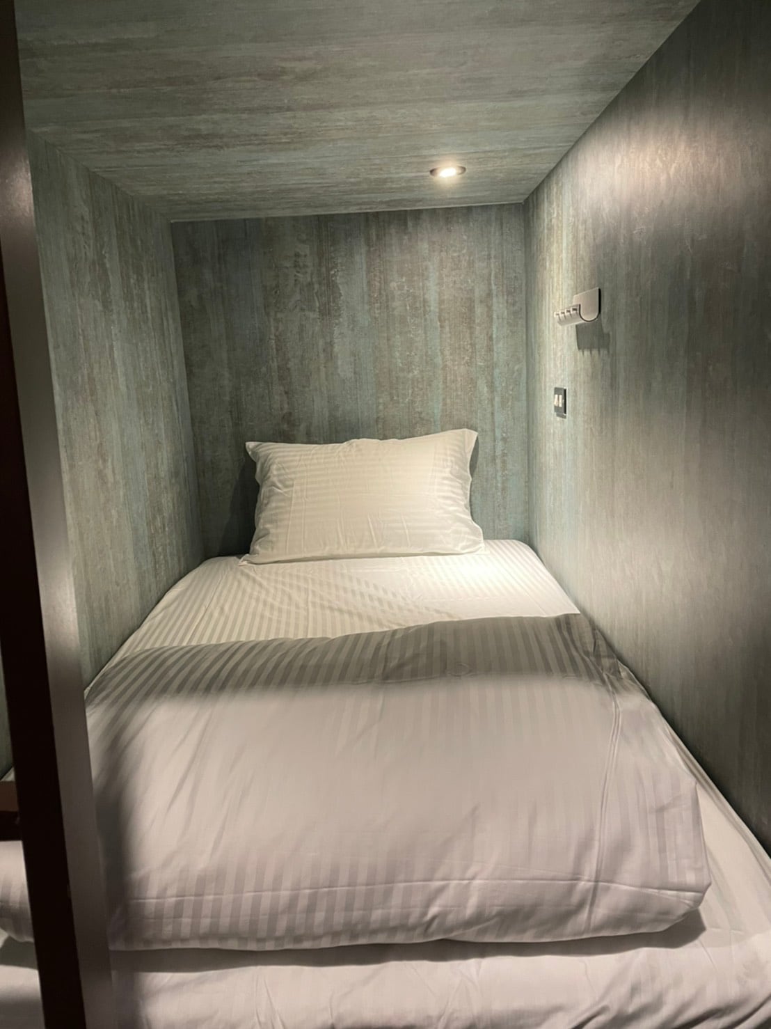 A compact sleeping space is shown, featuring a spacious bed with layered white bedding. A soft overhead light illuminates the interior, while the walls display a textured blue-grey finish. An outlet and reading light are accessible at the head of the bed.