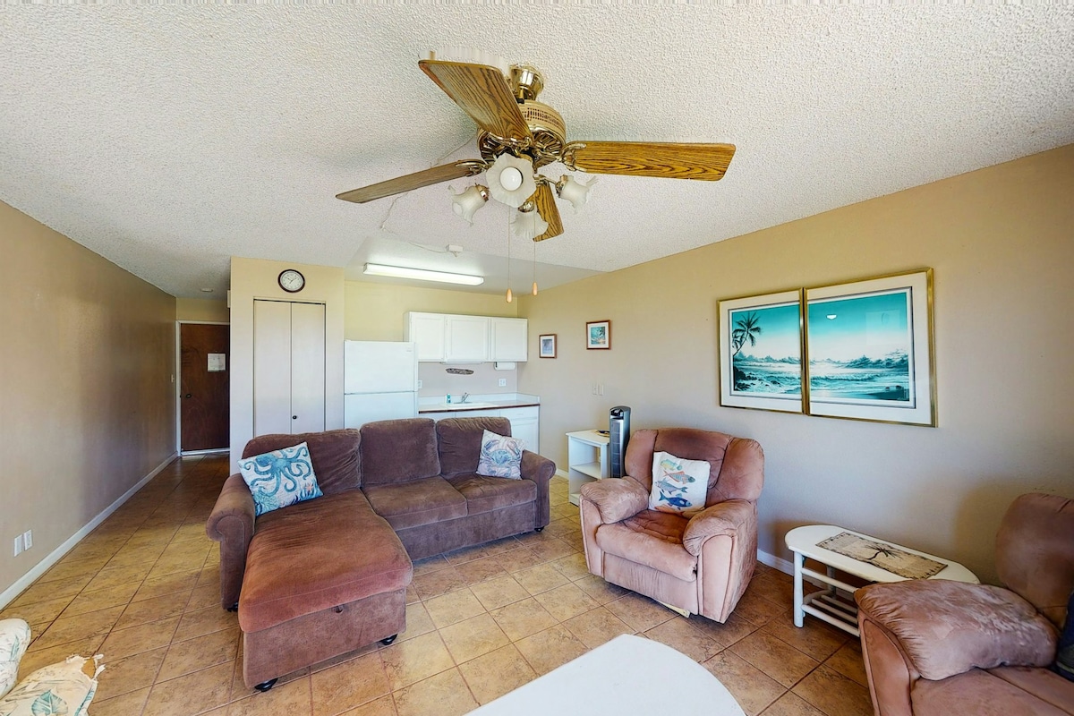 The spacious living area features a combination of cozy seating options, including a sectional sofa and armchairs. A ceiling fan hangs above, and a small dining table is visible in the background near the kitchen. Light-colored walls complement the tiled floor, creating an inviting atmosphere.