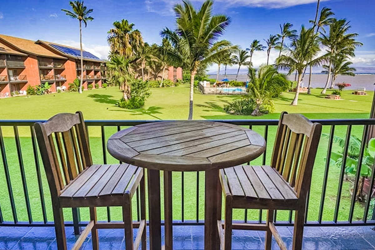 A lanai features a wooden table paired with two high stools, providing a view of lush greenery and swaying palm trees. The outdoor space overlooks a spacious lawn and an outdoor pool, with the Pacific Ocean visible in the distance.