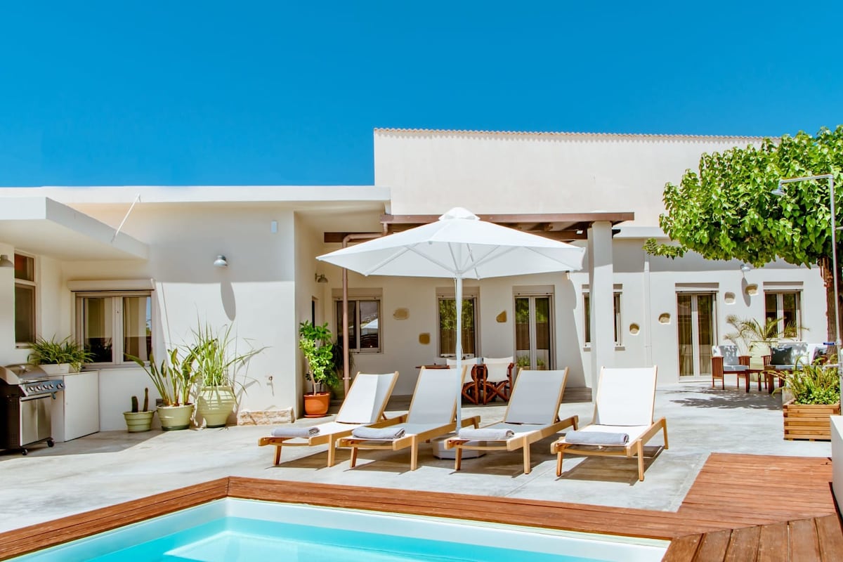 A modern outdoor space is showcased with a private swimming pool surrounded by comfortable loungers. An umbrella provides shade over the seating area. Vibrant plants are positioned around the space, enhancing the inviting atmosphere. The exterior features light-colored walls and large windows allowing natural light.