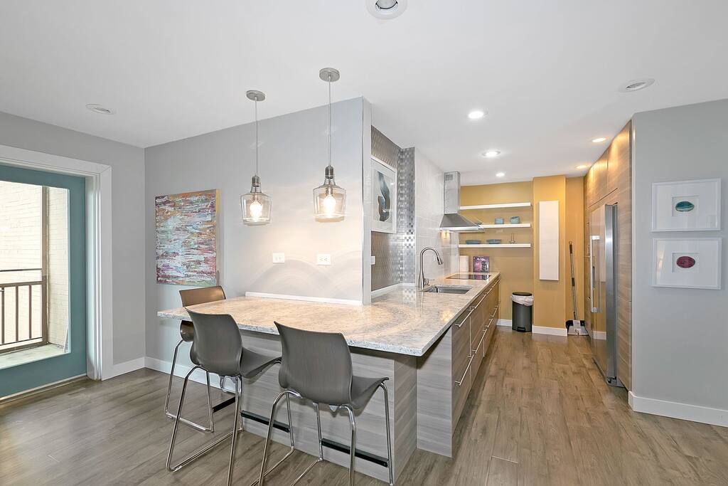 A modern kitchen area features a spacious island with two metal stools. Pendant lighting hangs above the countertop, which is adorned with granite. Cabinets and open shelving are visible, and a large stainless steel refrigerator is positioned against the wall.