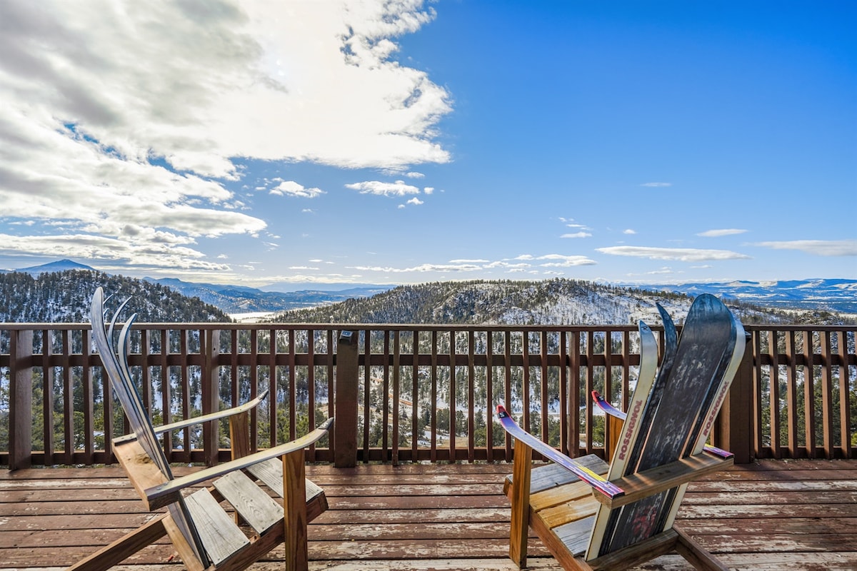 Wooden adirondack-style chairs are positioned on a spacious deck, offering expansive views of the surrounding snow-capped mountains and valleys. A clear blue sky is visible above, enhancing the serene outdoor setting.