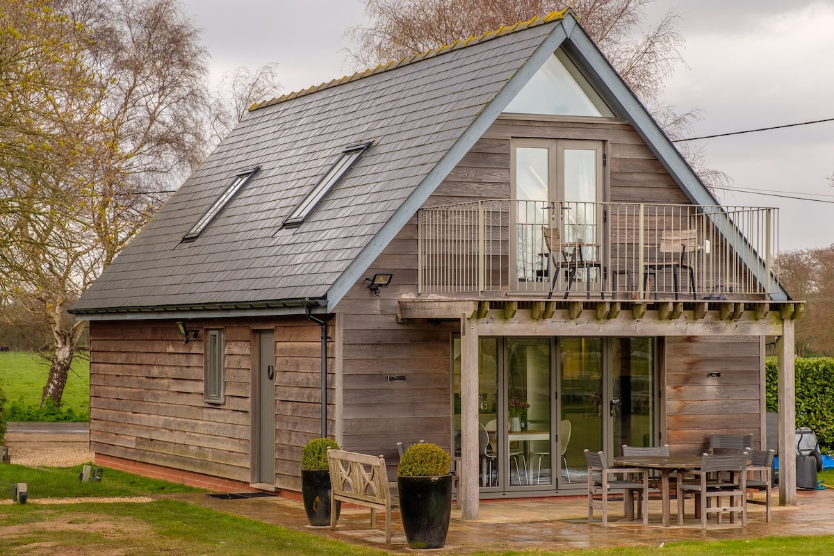 The exterior of a chalet-style building is showcased, featuring a modern wooden facade and a sloped roof. French doors lead to a spacious patio area with outdoor seating. A small balcony is visible on the upper level, offering views of the surrounding countryside.