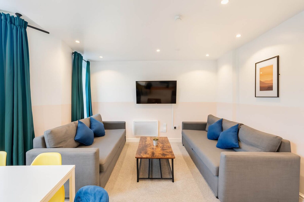 A modern living room is furnished with two gray sofas adorned with blue cushions, positioned across from a wooden coffee table. A flat-screen television is mounted on the wall, and bright curtains frame large windows, allowing ample natural light into the space.