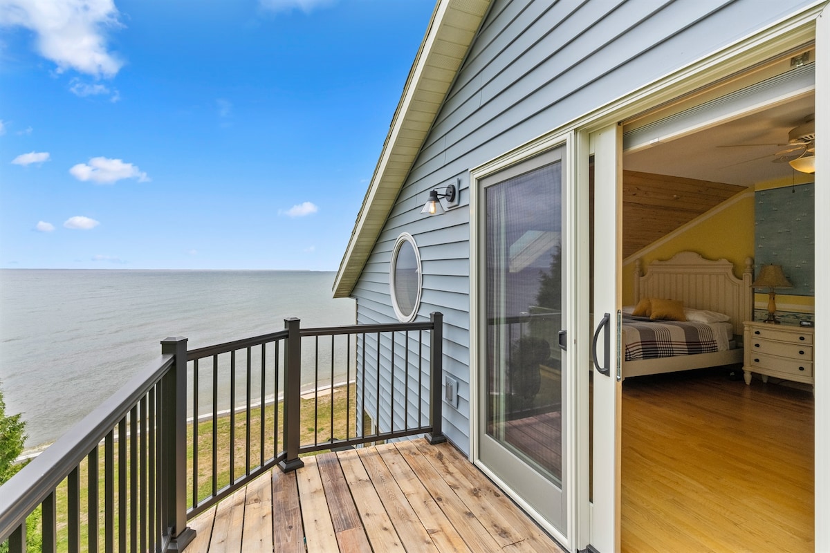 A serene balcony is visible, offering a clear view of the water with gentle waves. The space features a safety railing and is connected to a bright interior space that includes a cozy sleeping area adorned with light colors.
