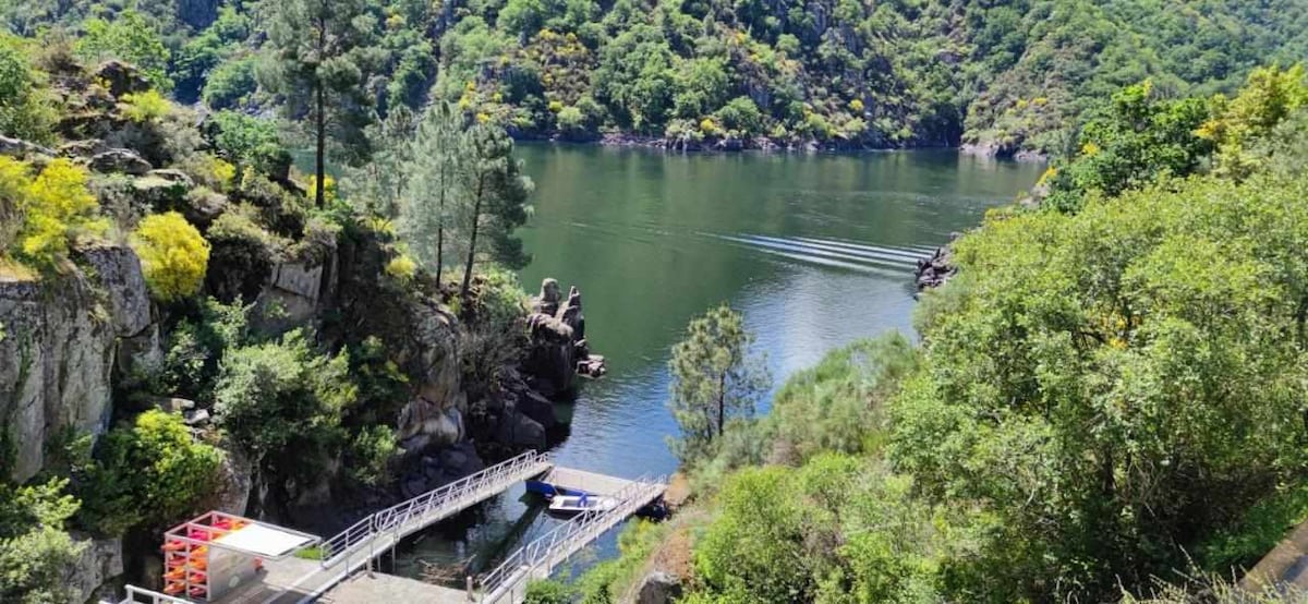 A serene river scene is framed by lush greenery and rocky landscapes. A small boat glides across the calm water, with a wooden dock extending into the river. Sunlight casts reflections on the surface, enhancing the natural beauty of the surroundings.