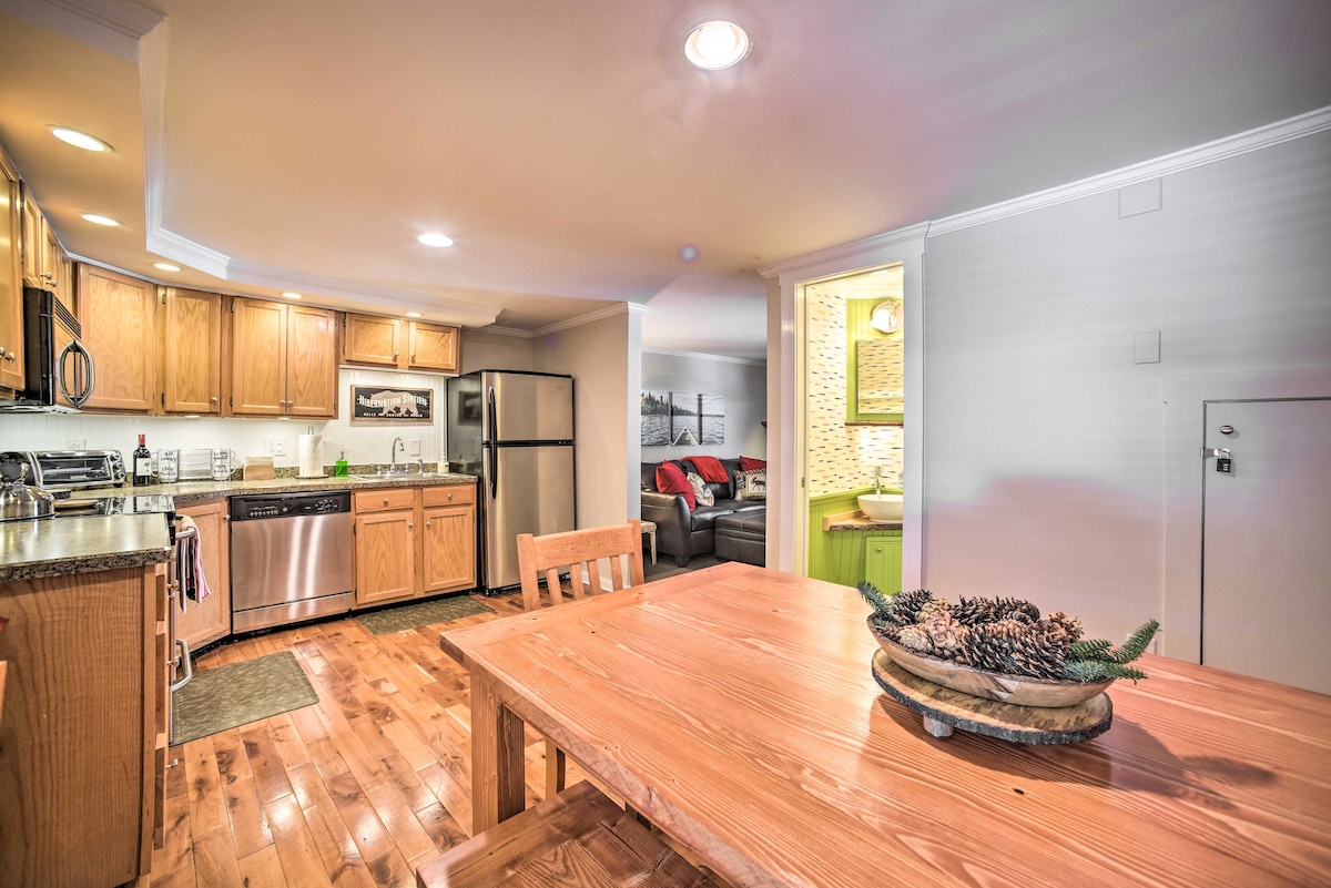 A well-equipped kitchen is visible featuring stainless steel appliances, including a refrigerator and dishwasher. A wooden dining table sits in the foreground, complemented by a decorative bowl containing pinecones. Soft lighting enhances the warm, wooden cabinetry and serene atmosphere.