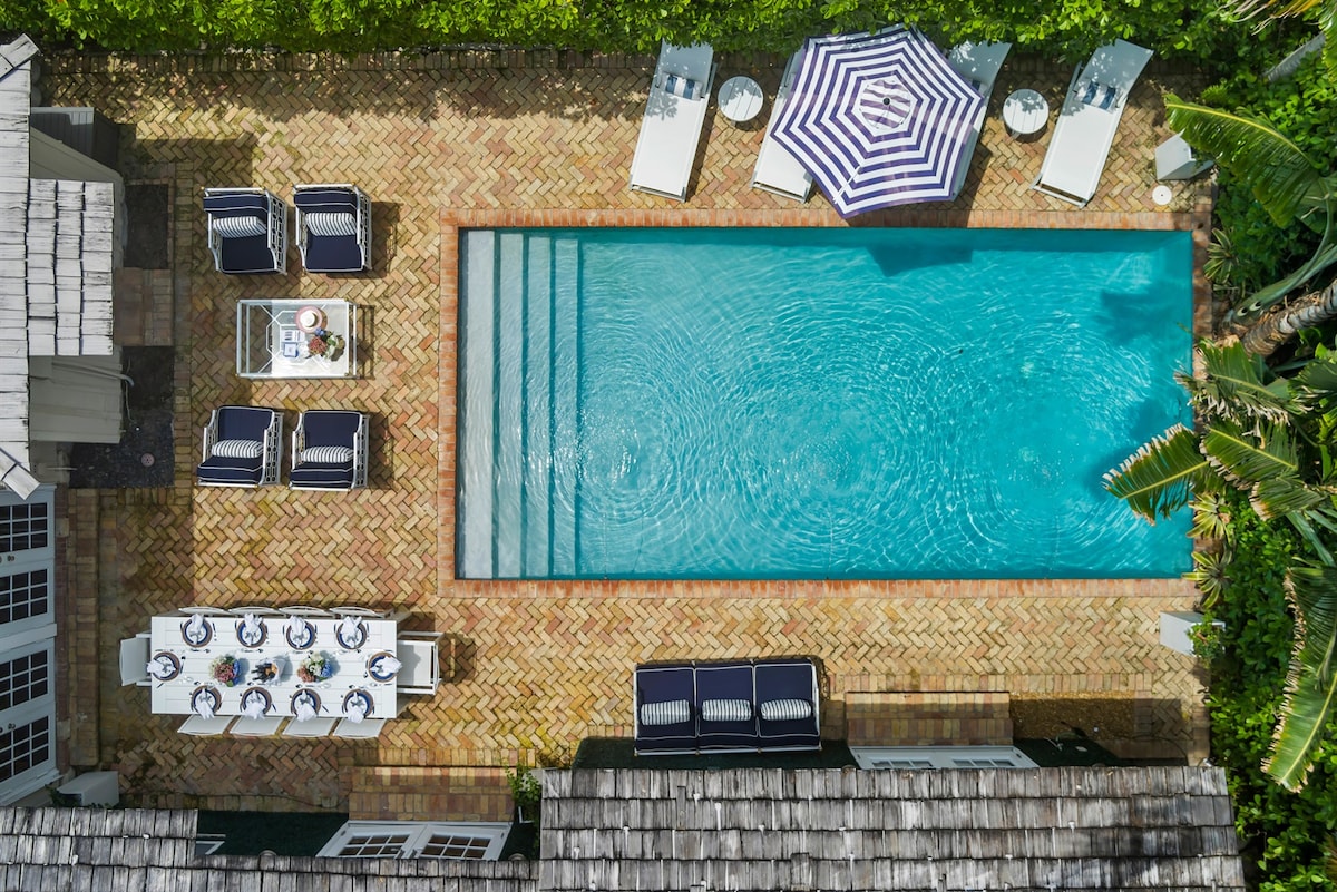 An aerial view captures a resort-style pool surrounded by lush greenery, offering lounge chairs and umbrellas. A spacious dining table is set nearby, providing ample seating options. The inviting water glistens under the sun, enhancing the outdoor living space's charm.