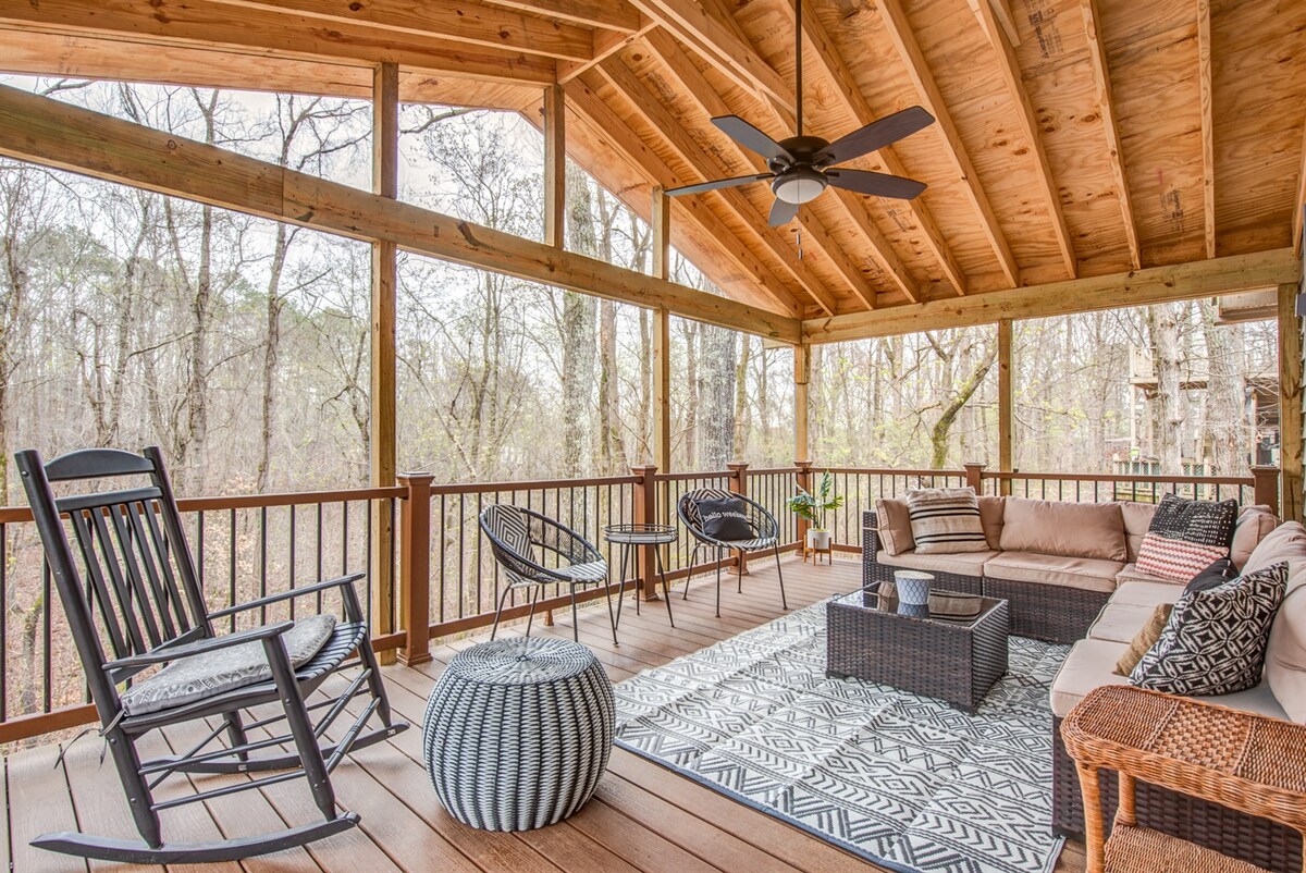 A spacious outdoor deck features a cozy seating arrangement with a large sectional couch and assorted chairs, providing ample space for relaxation. Natural wood beams support the high ceiling, and a coffee table sits at the center, surrounded by a serene forest backdrop.