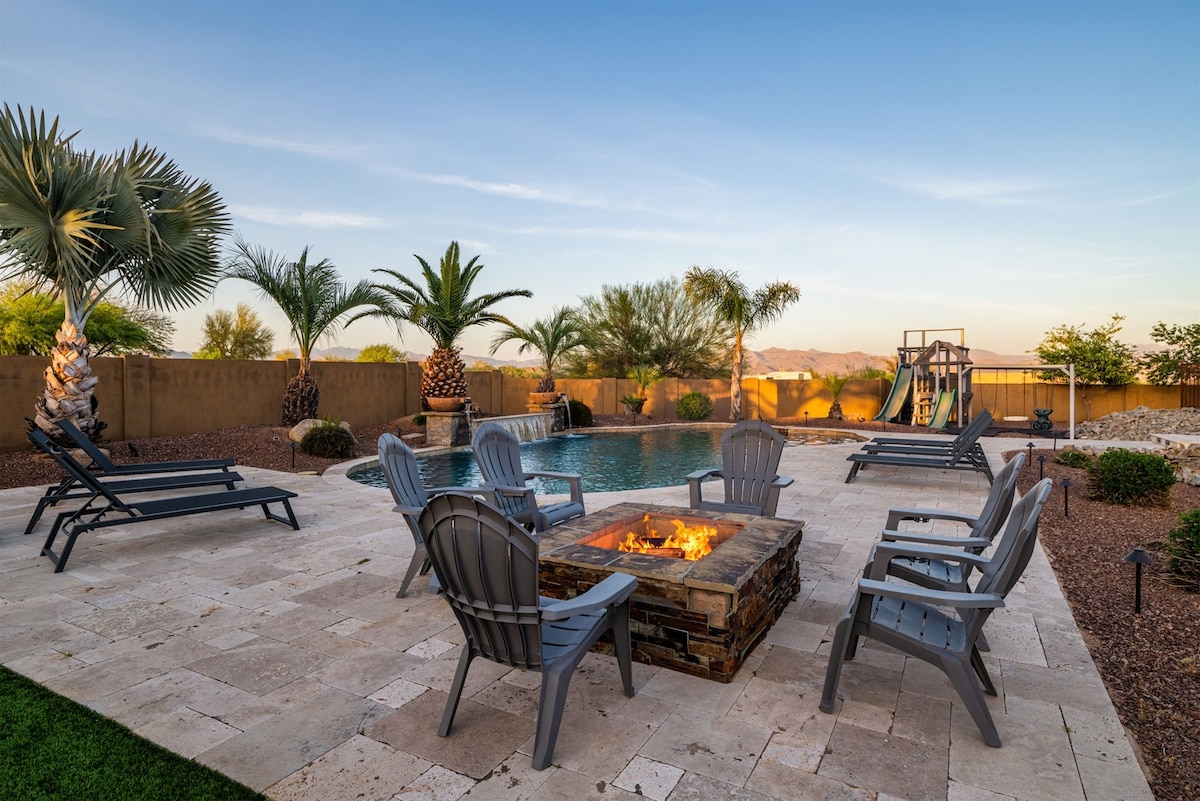An outdoor space is featured, showcasing a heated pool surrounded by a stone patio. Six gray lounge chairs are arranged near a stone fire pit, offering a gathering spot. Lush palm trees and desert landscaping are visible, with a slide structure in the background.