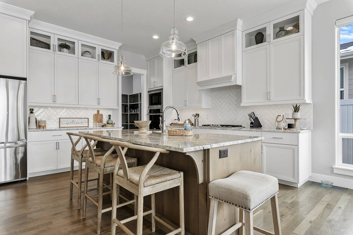 A modern kitchen showcases high-end stainless steel appliances, including a refrigerator and gas range. The spacious island, featuring seating for four, is topped with elegant granite. White cabinetry complements the herringbone backsplash, while ample natural light filters through large windows.