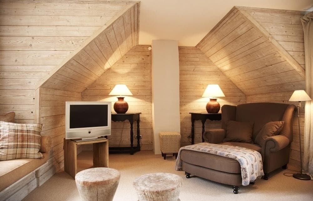A cozy living area features wooden walls with sloped ceilings, creating a warm atmosphere. A comfortable armchair is positioned next to a small side table, and a television sits on a simple cabinet. Soft lighting is provided by lamps on both sides of the room.