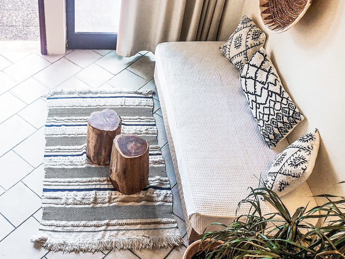 A cozy seating area features a light-colored, textured sofa adorned with patterned cushions. Natural wooden stools serve as a coffee table, positioned on a striped area rug. Soft natural light enters from a nearby window, highlighting the simplicity of the space.