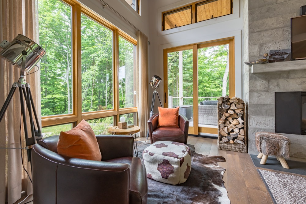 A bright living space features large windows that showcase views of the surrounding forest. Two leather armchairs with orange pillows are positioned near a side table. A decorative pouf rests on a patterned rug, while a wood stack is neatly arranged by the fireplace.