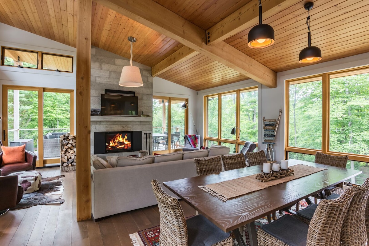 A spacious living and dining area features large windows that provide a view of the natural surroundings. A stone fireplace serves as a focal point, while a wooden dining table surrounded by woven chairs accommodates guests. A warm wooden ceiling enhances the cozy ambiance.