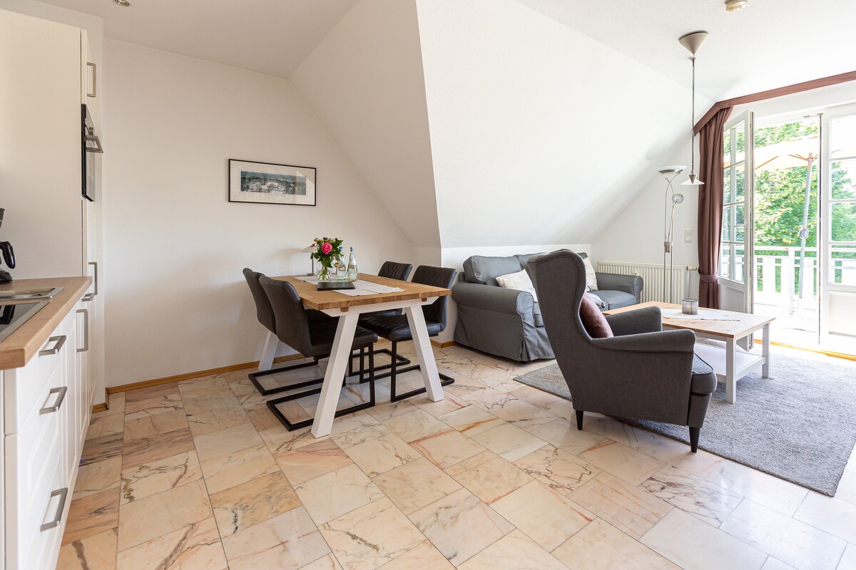 An open living area features a dining table with six chairs and a cozy seating area with a combination of a sofa and armchairs. Sliding glass doors allow natural light and provide access to a balcony overlooking greenery. Marble flooring adds a touch of elegance.