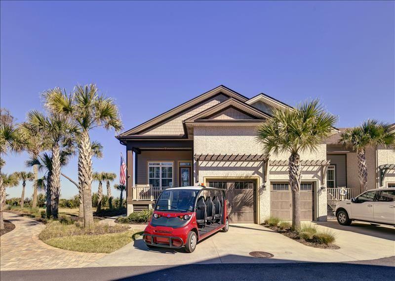 A cottage-style townhome is showcased with a well-maintained exterior featuring palm trees and a wide driveway. A vibrant six-seater electric cart is parked in front, ready for exploration, while the charming architecture highlights the welcoming atmosphere of the beach community.