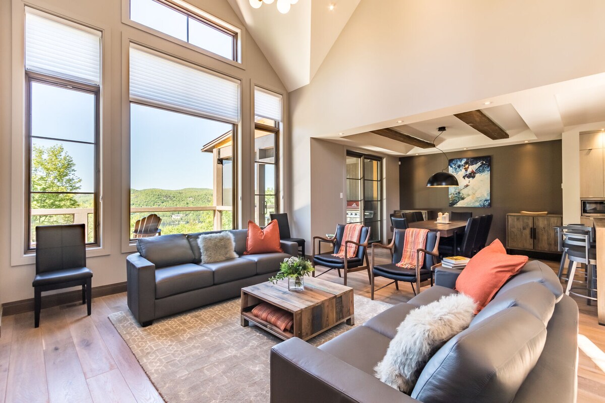 A spacious living area is illuminated by natural light streaming through large windows, offering views of the surrounding landscape. Comfortable seating is arranged around a coffee table, with a dining area in the background. Modern accents are presented throughout the design.