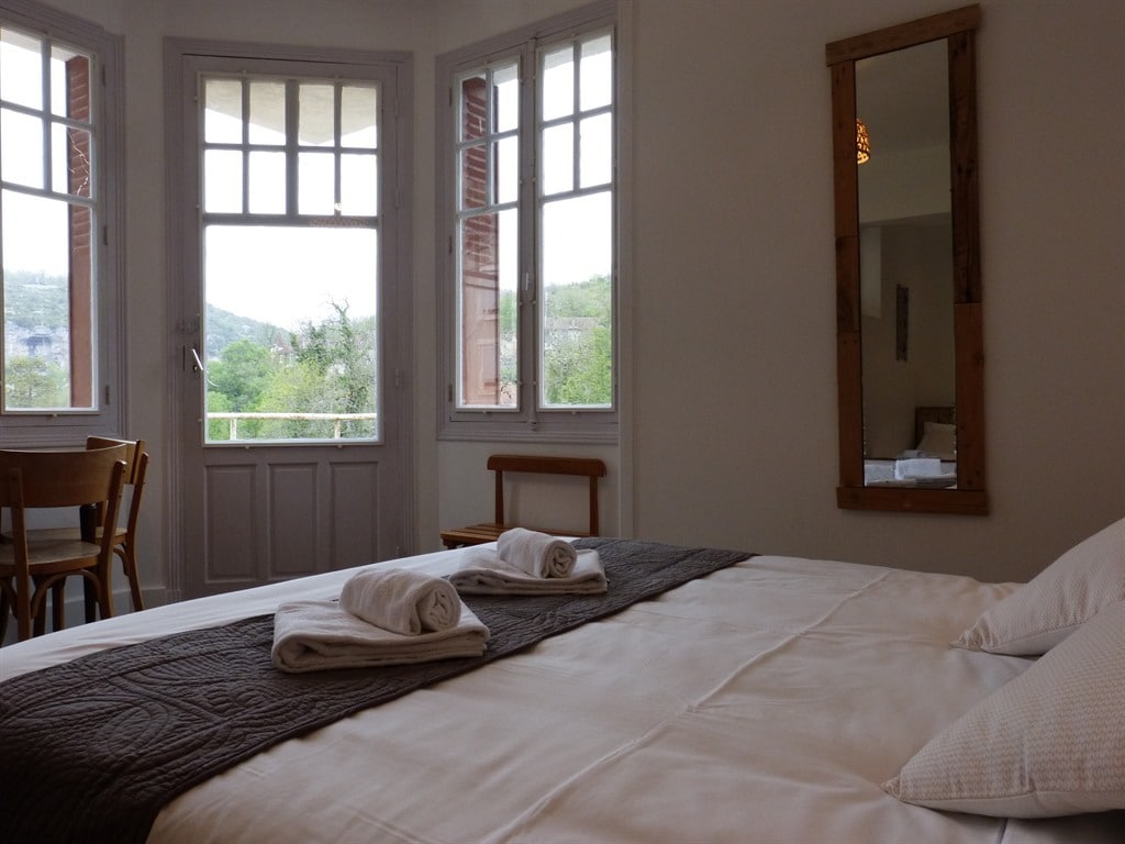 A spacious room features a large double bed with neatly arranged towels. Natural light floods in through multiple windows, offering views of the surrounding greenery. A simple wooden chair and table are included, complemented by a framed mirror on the wall.