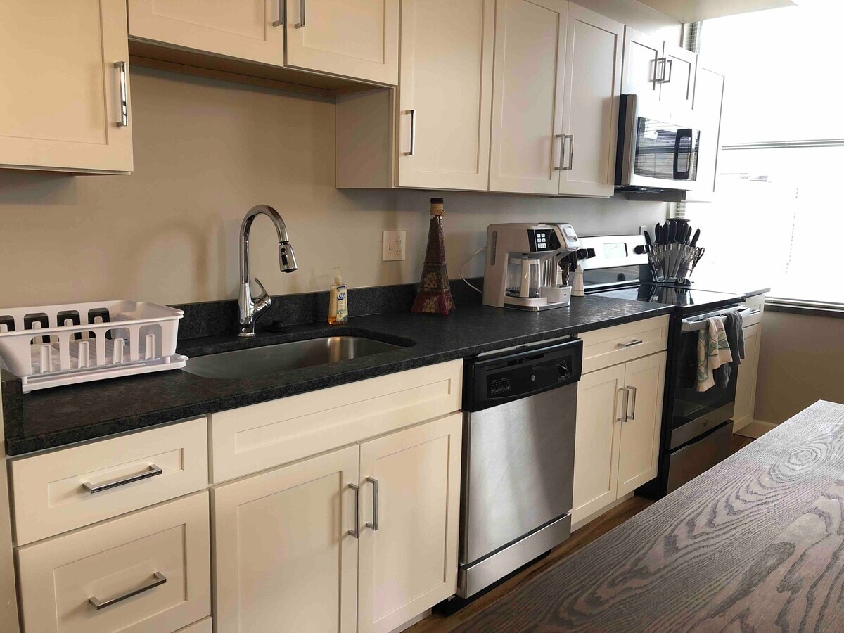 A modern kitchen is equipped with light cabinetry and dark countertops. Stainless steel appliances include a microwave, dishwasher, and coffee maker. A sink is positioned beneath a window, and kitchen utensils are neatly organized in a holder. A dining table with a wood finish is located nearby.