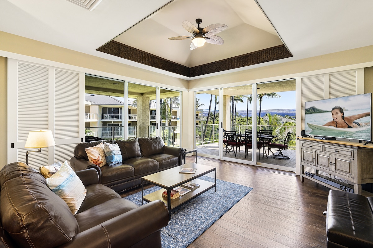 The living area is furnished with a brown leather sofa set and a glass-topped coffee table, complemented by a colorful area rug. A flat-screen TV is positioned against a side cabinet. Large sliding glass doors provide natural light and scenery views.