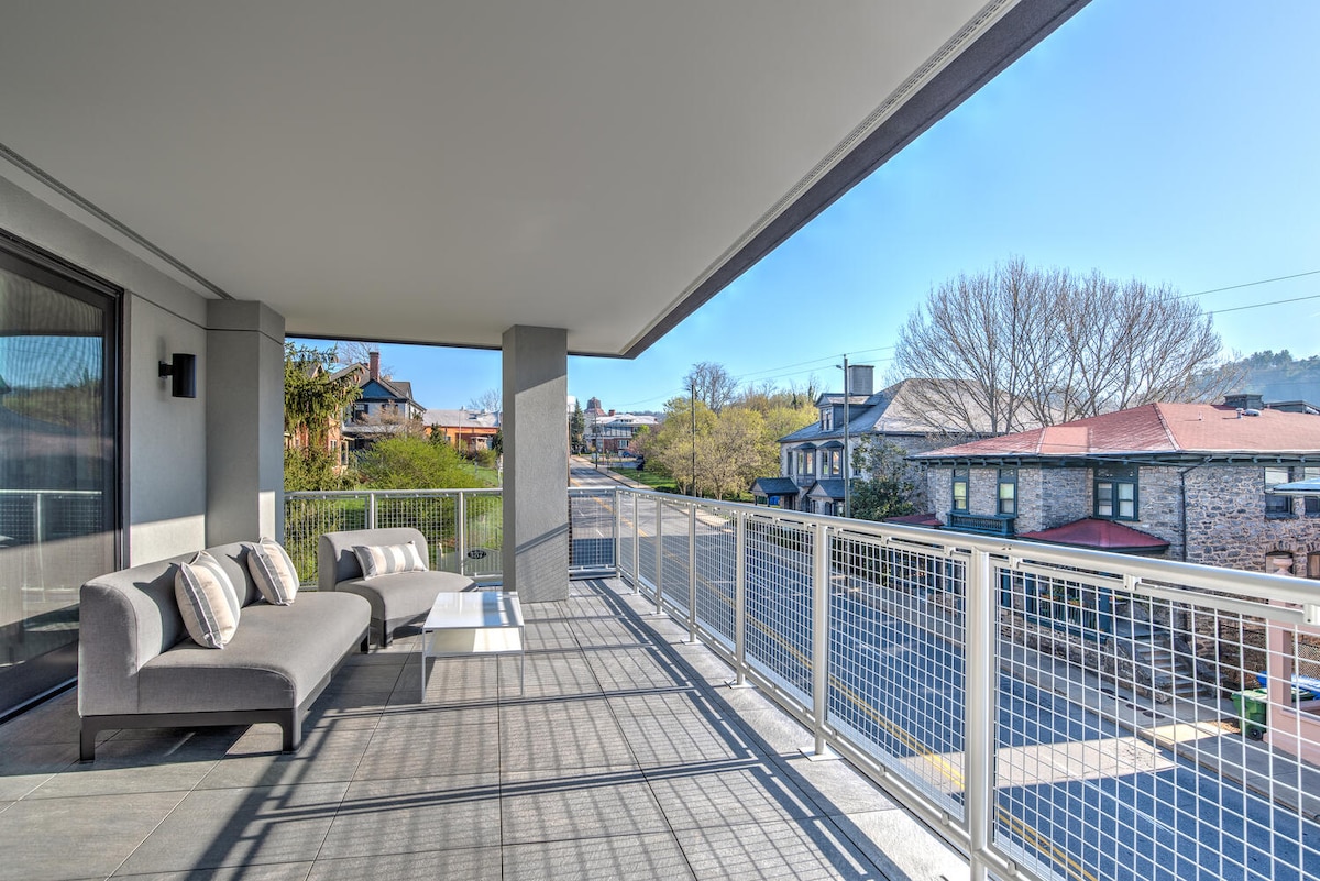 A spacious outdoor balcony features a modern gray sofa with decorative pillows and a low coffee table. The surrounding railings provide unobstructed views of the neighborhood, with sunlight illuminating the area and trees visible in the distance.