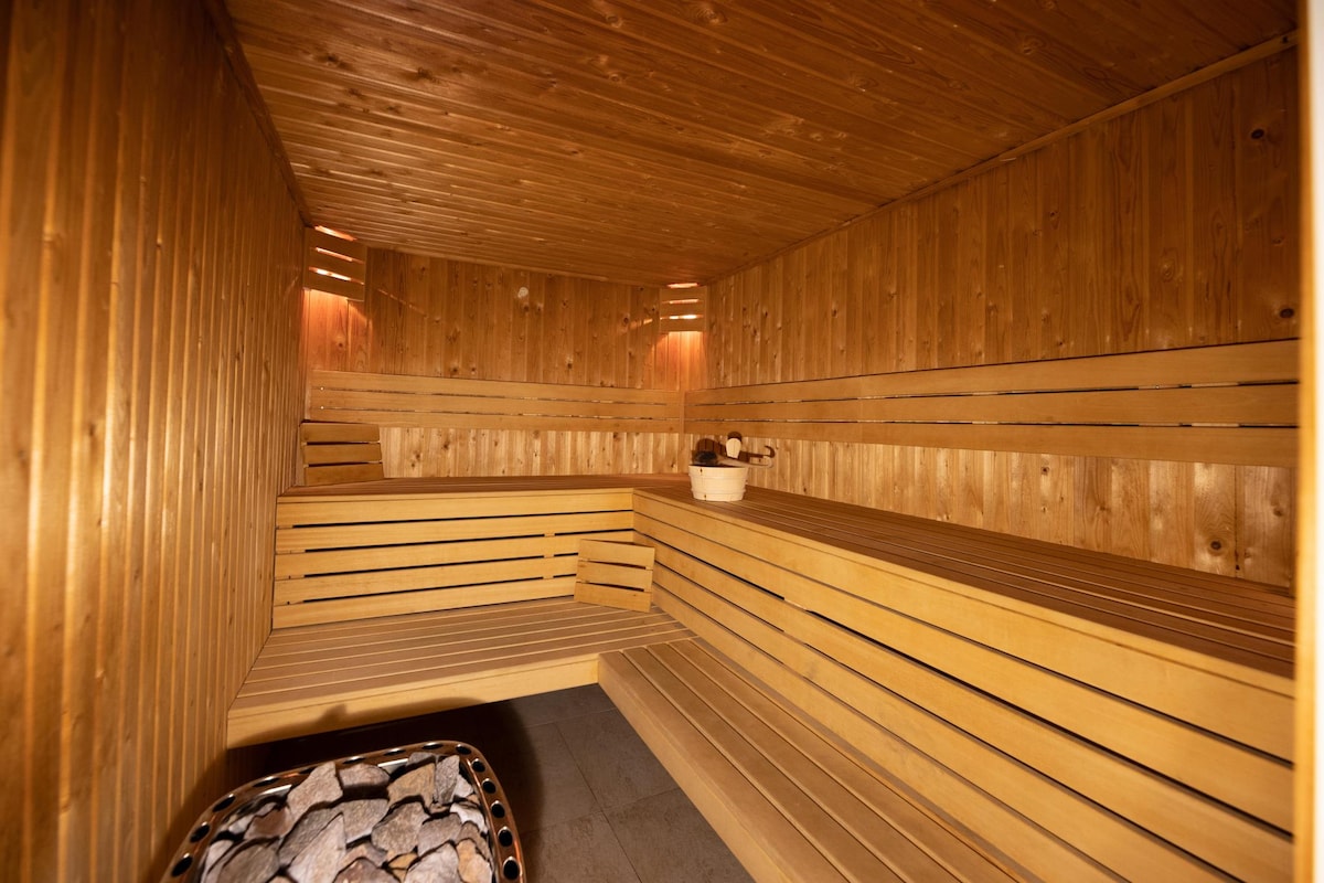 A wooden sauna is presented with multiple bench seating areas, designed for relaxation. Natural wood paneling accentuates the tranquil environment, while a stone heater is visible, ready to provide warmth.