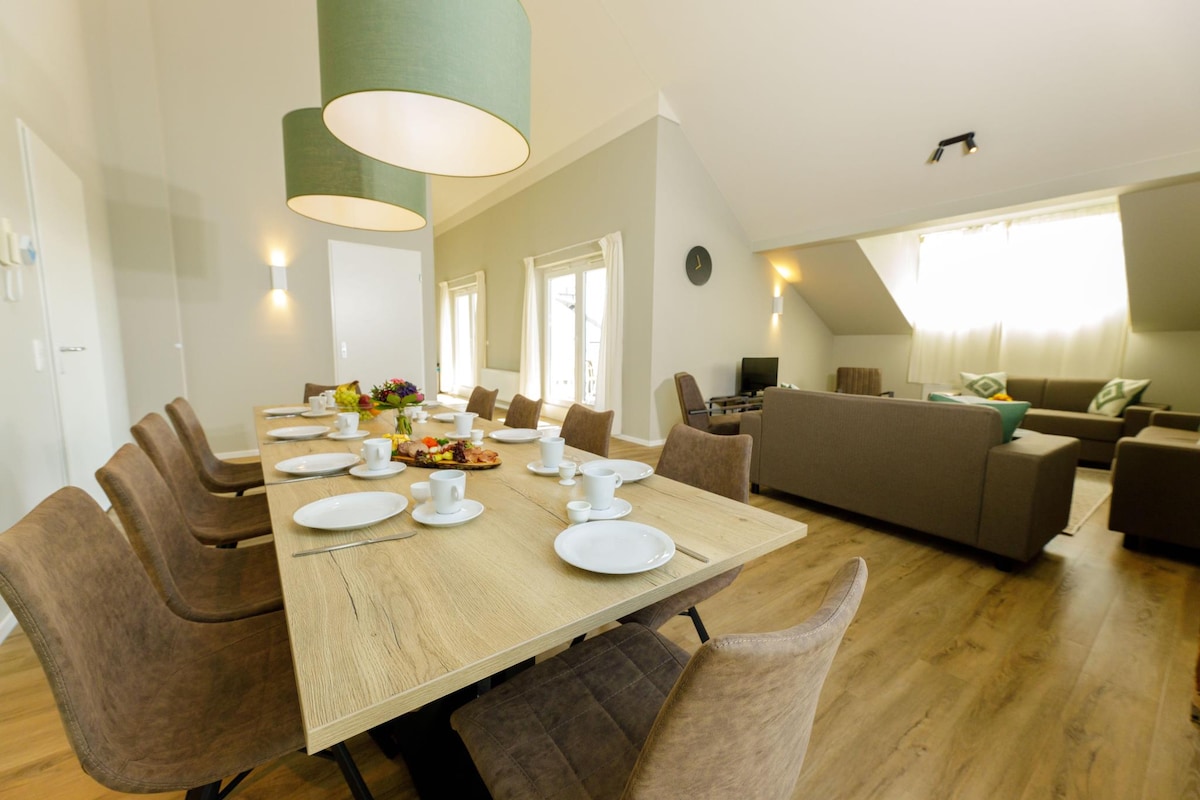 A spacious dining area is equipped with a large wooden table set for breakfast, featuring plates and cups. Soft green pendant lights illuminate the room, which is enhanced by large windows and a cozy seating area visible in the background.